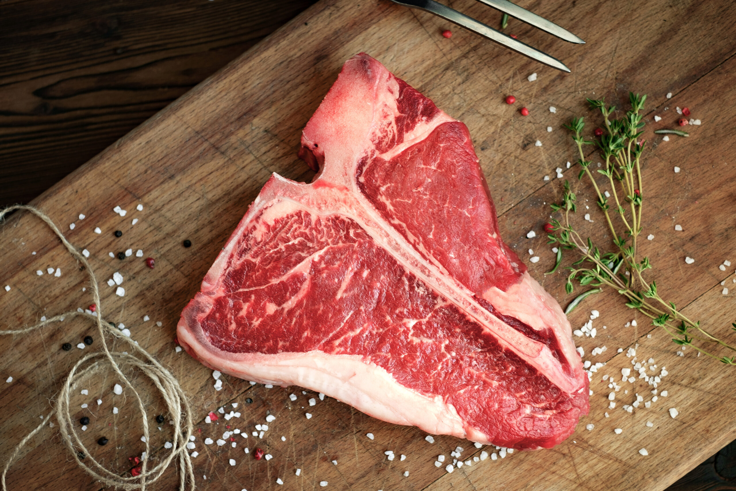 Fresh T-bone steak on a wooden cutting board, surrounded by coarse sea salt, black and pink peppercorns, and sprigs of thyme, ideal for grilling or roasting.